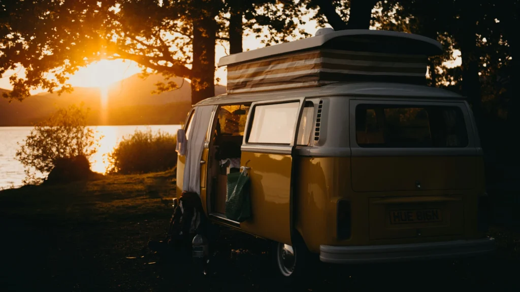 Ein alter VW-Camper steht in der Dämmerung unter Bäumen am Ufer eines Sees. Vor der geöffneten Seitentür ist im Gegenlicht schemenhaft ein Mensch zu erkennen.