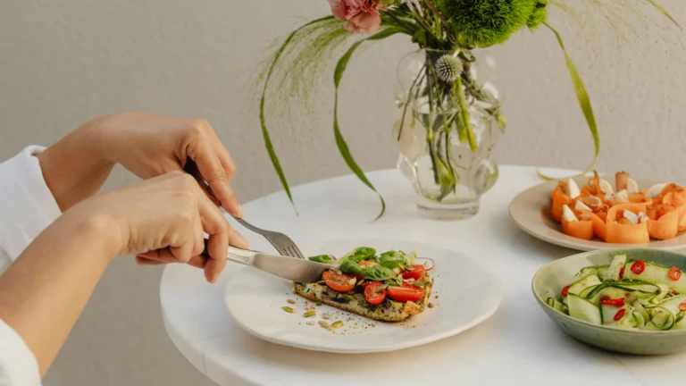 Auf einem Tisch stehen eine Blumenvase, zwei kleine und ein großer Teller mit sommerlichen Speisen. Von links ragen zwei Hände ins Bild, die mit Messer und Gabel ein Stück einer Bruscchetta abschneiden.