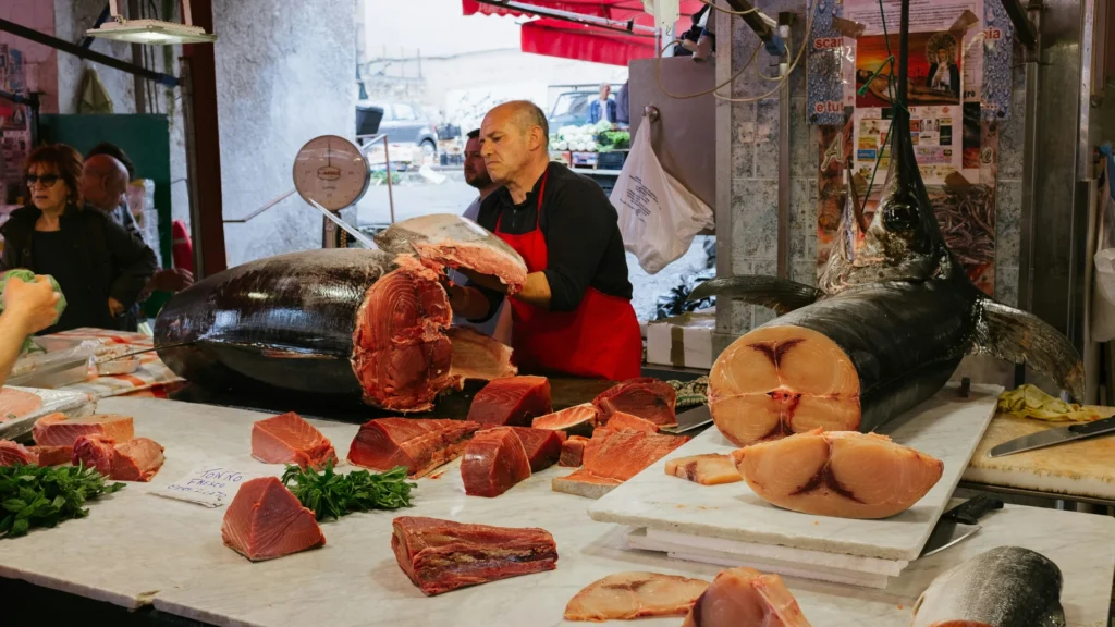 Ein kahlköpfiger Mann mit schwarzem Hemd und roter Schütze filetiert in einer Fischhandlung einen Thunfisch. Seitlich sind Kundinnen und Kunden zu sehen.