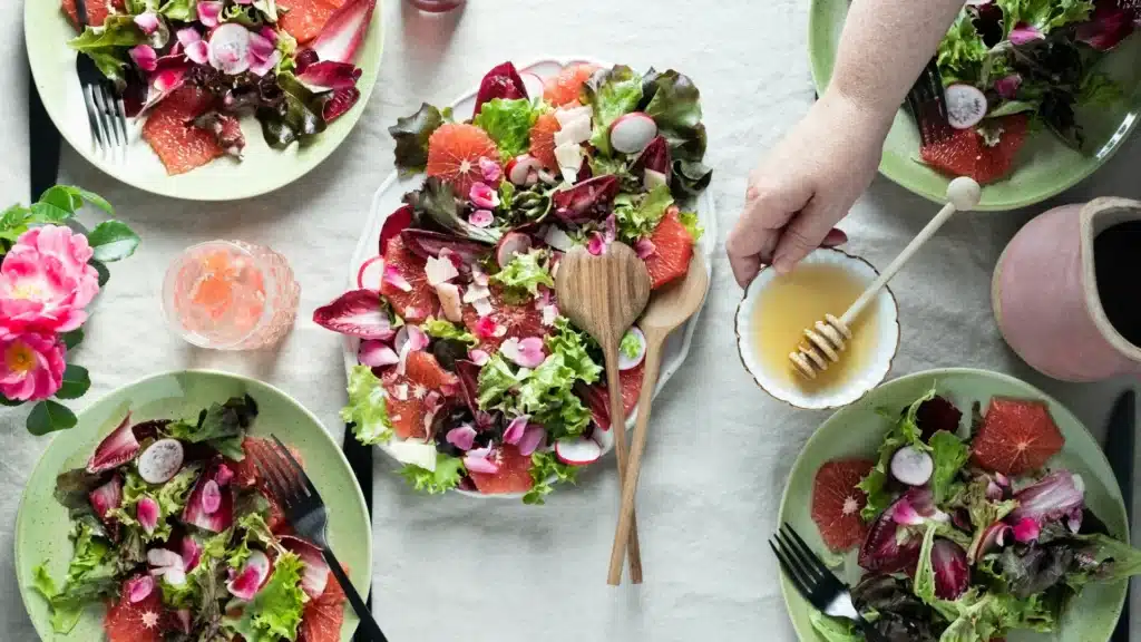 Ein Tisch mit weißer Decke. Auf ihm stehen mehrere Teller mit Salat. Eine Hand reicht einie kleine Schüssel mit Dressing ins Bild.