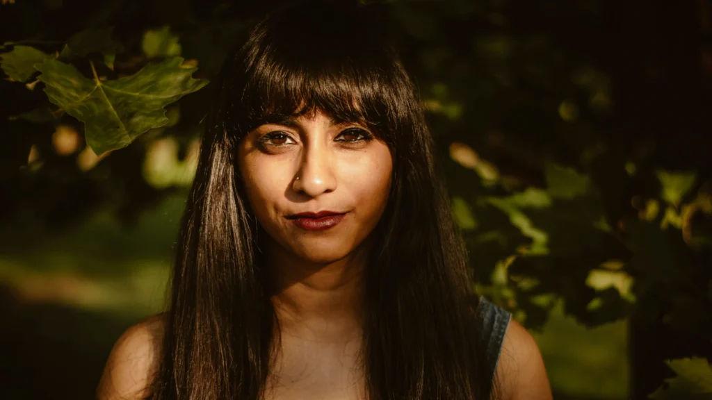 Eine Frau mit braunen langen Haaren steht im Schatten eines Obstbaums. Sie blickt lächelnd in die Kamera. Auf ihr Gesicht fällt ein Sonnenstrahl.