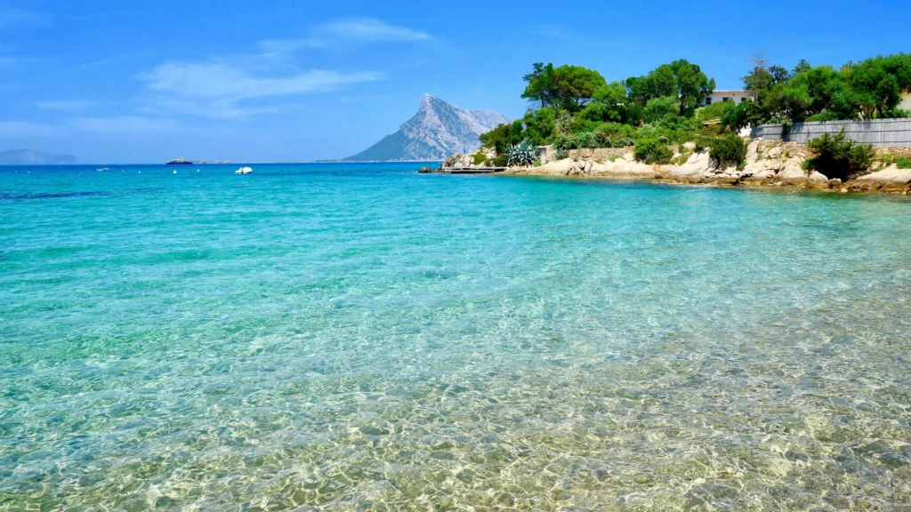 Am Strand. Vorn azurblaues Wasser über Sand, rechts die Küstenlinie, weiter hinten Boote und ein Berg.