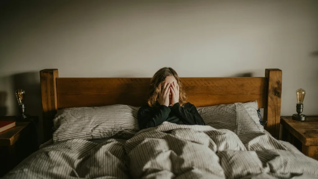 Eine Frau liegt in einem Hozbett mit schwarz-weiß-gestreifter Bettwäsche. Sie schlägt ihre Hände vors Gesicht.