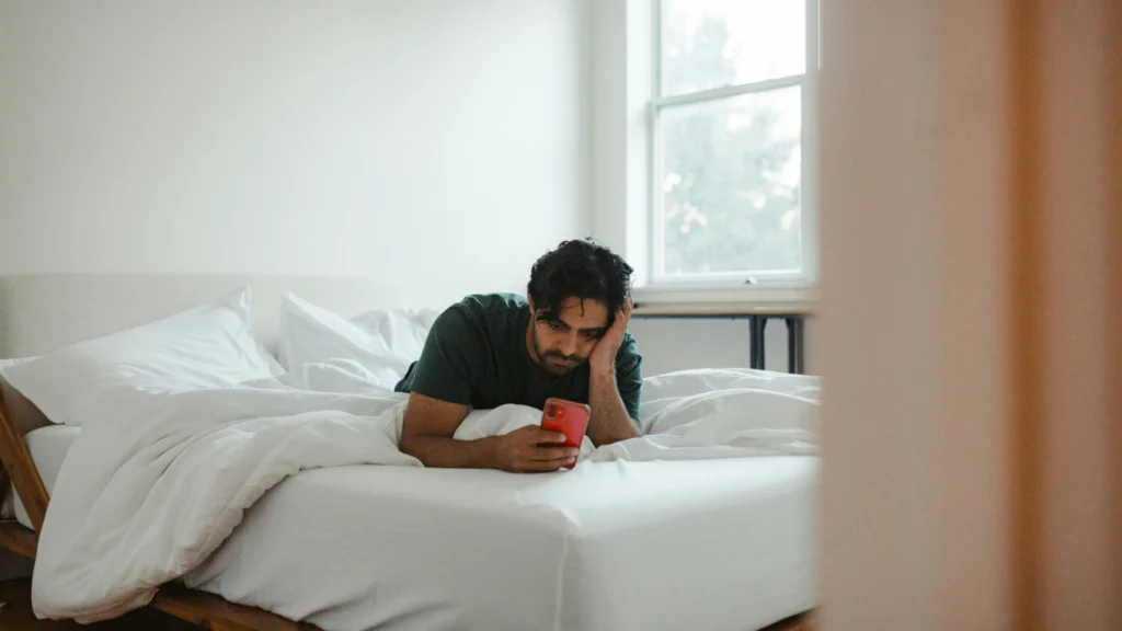 Ein Mann liegt bäuchlings im Bett. Er blickt grübelnd auf sein Smartphone.
