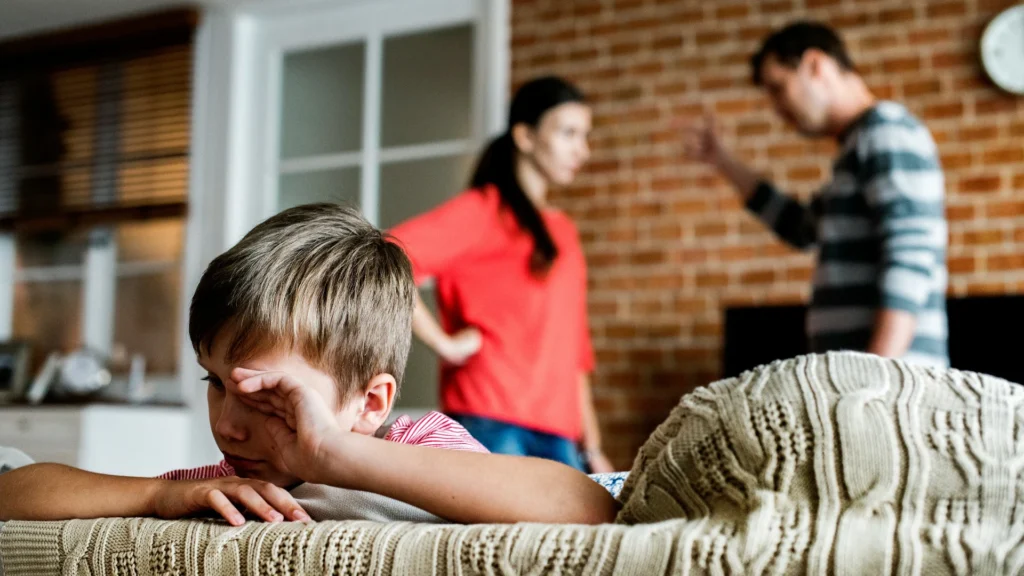 Eine Wohnung. Im Bildhintergrund streiten sich ein Mann und eine Frau. Vorn sitzt ein trauriger Junge auf einem Sofa.