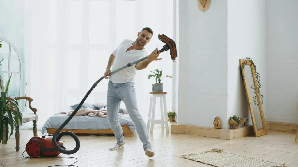 Ein Zimmer mit weißen Wänden und einem hellen Dielenboden. Im Raum befinden sich eine Grünpflanze, ein Stuhl, zwei Spiegel, ein Futonbett und verschiedene Dekogegenstände. Davor steht ein Mann, der mit dem Rohr seines Bodenstaubsaugers posiert.