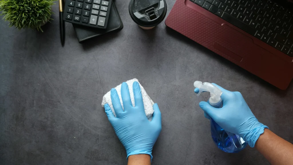 Zwei Hände in blauen Gummihandsschuhen. Die linke hält ein Putztuch, die rechte eine Sprühflasche über eine schwarze Platte, auf der Tastatur, Kaffeebecher, REchner, Notizbuch und Kugelschreiber liegen.