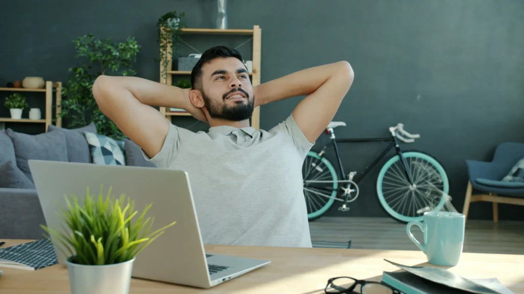 Ein Mann leht sich an seinem Schreibtisch gemütlich zurück. Er hart seine Hände hinter dem Kopf verschränkt. Auf dem Tisch steht ein Laptop und eine Kaffeetasse, an der Wand hinter ihm Regale, Grünpflanzen, ein Sessel und ein Fahrrad.