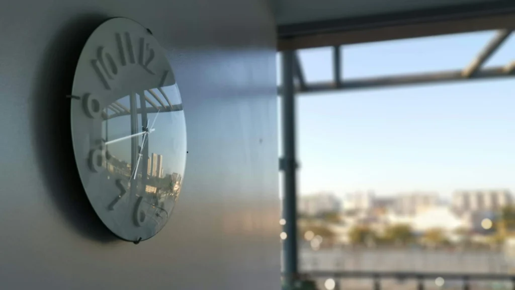 Links an der Wand hängt eine Uhr. An ihr vorbei geht der Blick über einen Balkon auf die verschwommene Silhoutte einer Stadt.