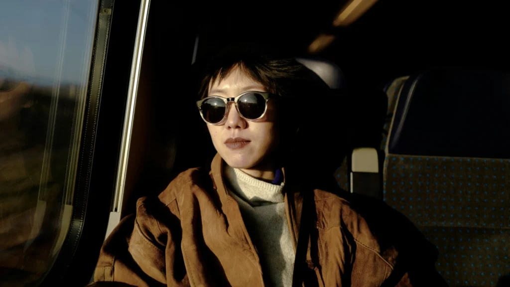 Eine Frau mit braunen kurzen Haaren sitzt in einem Bus. Sie trägt eine Sonnenbrille, eine braune Lederjacke und einen beigen Pullover. Durchs Fenster wird sie von der Sonne beschienen.