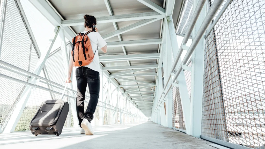 Ein Mann mit braunem Dutt telefoniert, während er auf einer Brücke mit Rucksack und Rollkoffer unterwegs ist. Er trägt ein weißes T-Shirt und schwarze Jeans.