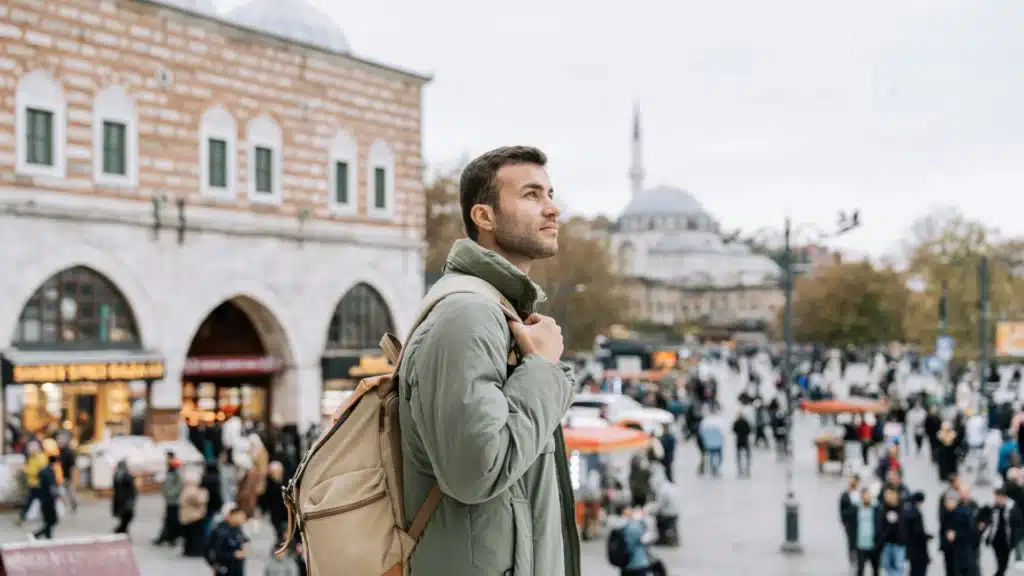 Ein bärtiger Mann mit Ruchsack steht auf einem belebten Platz. Im Hintergrund sind ein Gebäude mit Bogengängen und eine Moschee zu sehen.