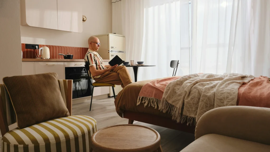 Ein kahlköpfiger Mann sitzt lesend am Esstisch seines Apartments. Auf dem Bild sind ebenfalls eine Couch mit Beistelltisch, ein Bett und eine Küchenzeile zu erkennen.