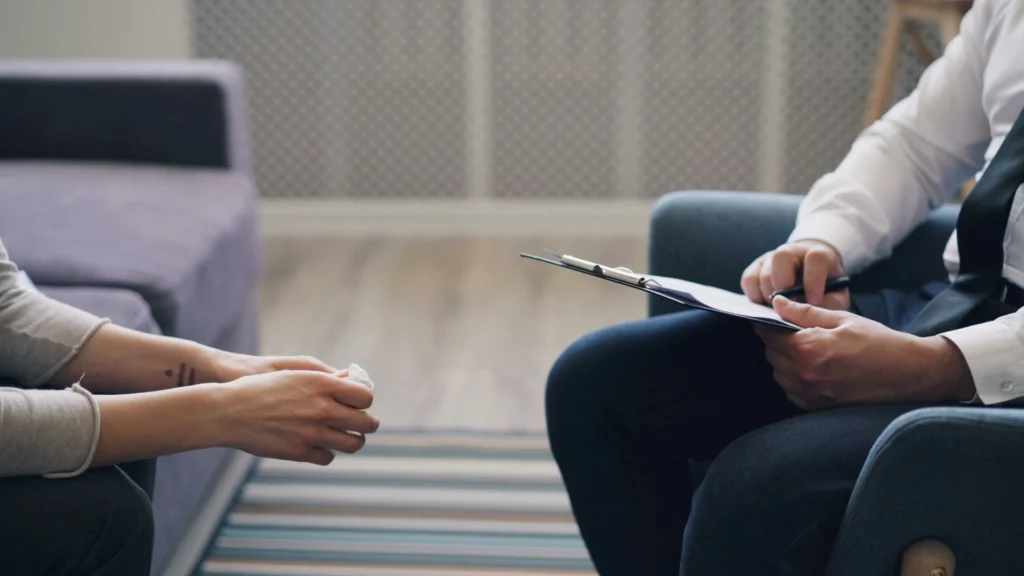 Zwei Menschen sitzen sich auf Sofas gegenüber. Von ihnen sind nur Hände und Knie zu sehen. Die linke Person hat die Hände gefaltet, die rechte Person macht Notizen in ein Buch.