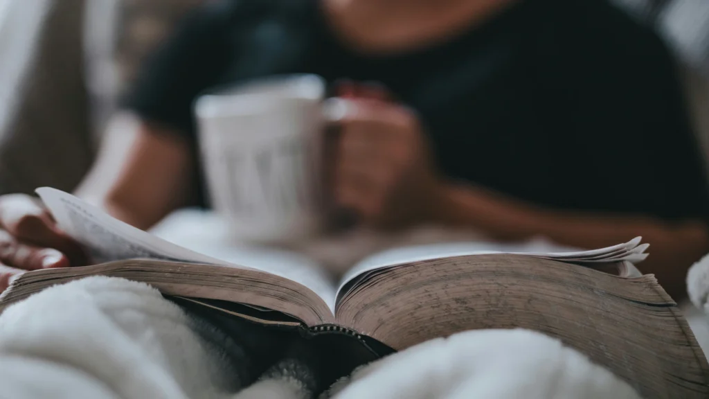 Eine Person liegt mit zugedeckten Beinen auf dem Sofa. Auf ihrem Schoß liegt ein Buch, darauf die rechte Hand. In der linken hält sie eine Tasse.