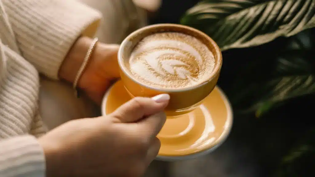 Die Hände einer Frau halten eine Cappuccinotasse, in deren Schaum sich ein Blatt abzeichnet.