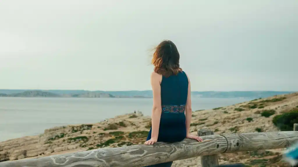 Eine Frau in einem blauen Kleid lehnt mit dem Rücken zur Kamera auf einem Holzzaun an einem Sandstrand. Sie blickt hinaus aufs Meer.