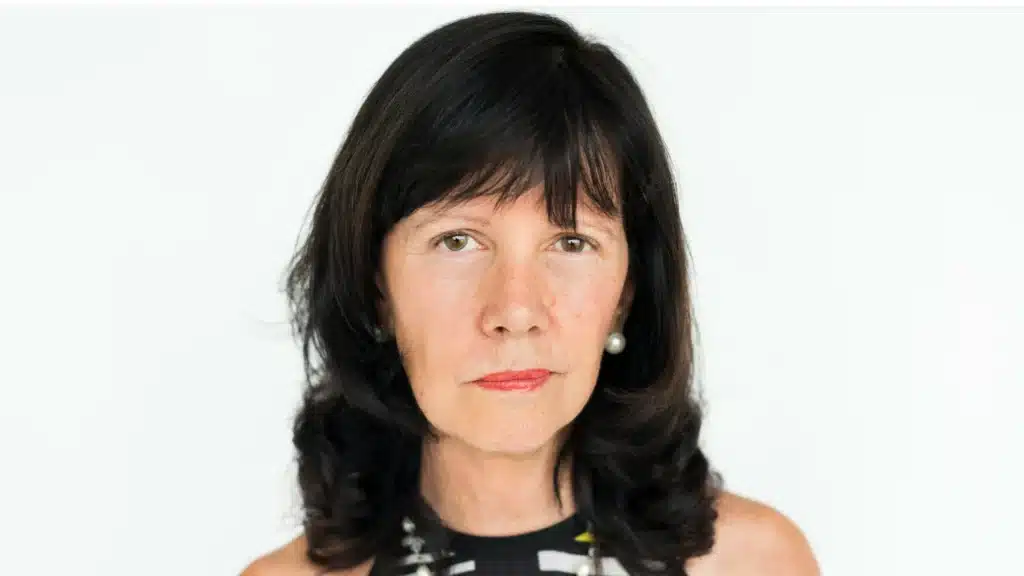 Eine Frau mit halblangen dunklen Haaren steht vor einer weißen Wand. Sie blickt direkt in die Kamera.