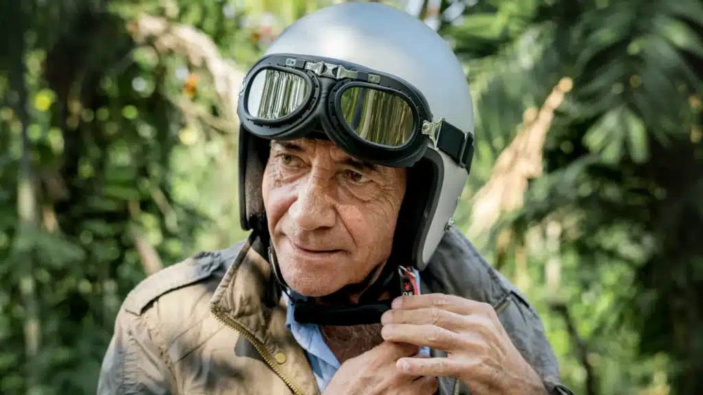 Ein Mann mittleren Alters trägt Motorradhelm und -brille. Seine Hände öffnen den Verschluss des Helms.