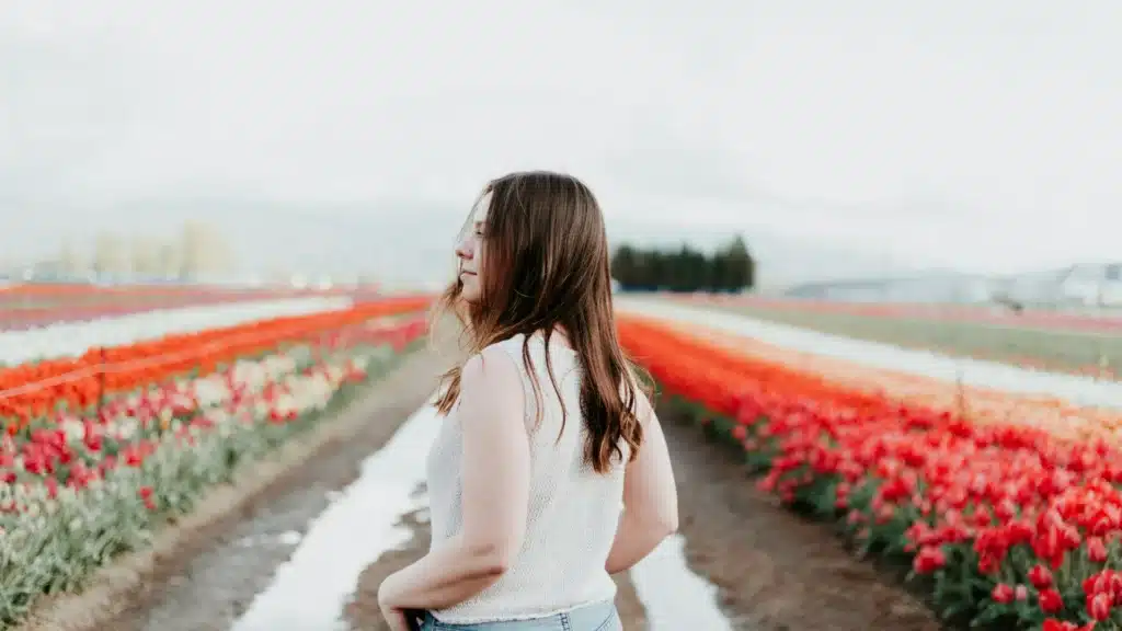 Eine Frau mit langen braunen Haaren steht mit dem Rücken zur Kamera in einem Tulpenfeld. Sie trägt ein weißes Stricktop und blickt lächelnd nach links.
