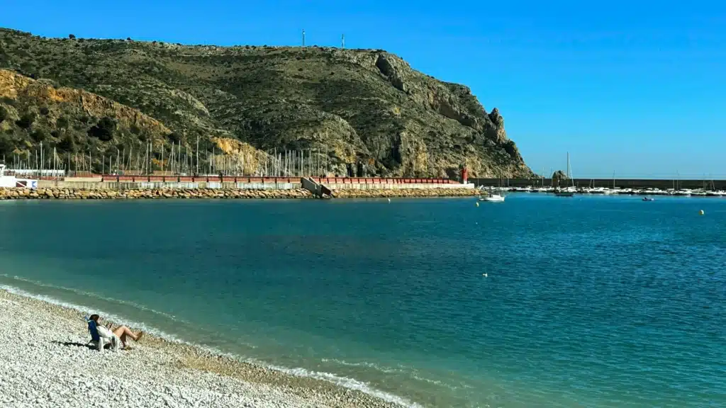 Eine Frau sitzt lesend allein in einem Liegestuhl am Strand. Im Bildhintergrund sind eine Mole, Boote und Hügel zu erkennen.