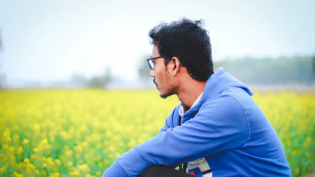 Ein Mann mit dunklen Haaren und Vollbart sitzt seitlich zur Kamera in einer Blumenwiese. Er trägt eine schwarze Brille und einen blauen Hoodie.