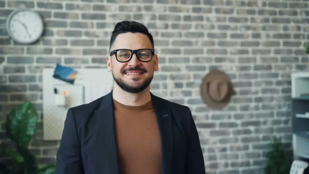 Ein Mann mit dunklen Haaren und Vollbart blickt lächelnd in die Kamera. Er trägt eine schwarze Brille, ein blaues Sakko und einen braunen Pullover. Hinter ihm ist eine Ziegelwand zu sehen, an der eine Uhr, ein Whiteboard und ein Hut hängen.