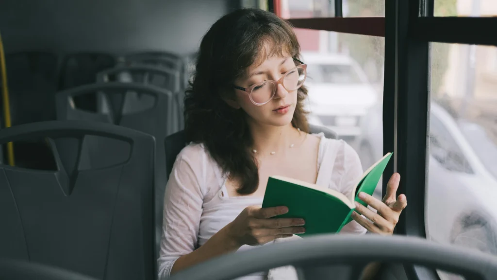 Eine junge Frau mit braunen langen Haaren und einer Brille liest in einem Bus ein Buch.