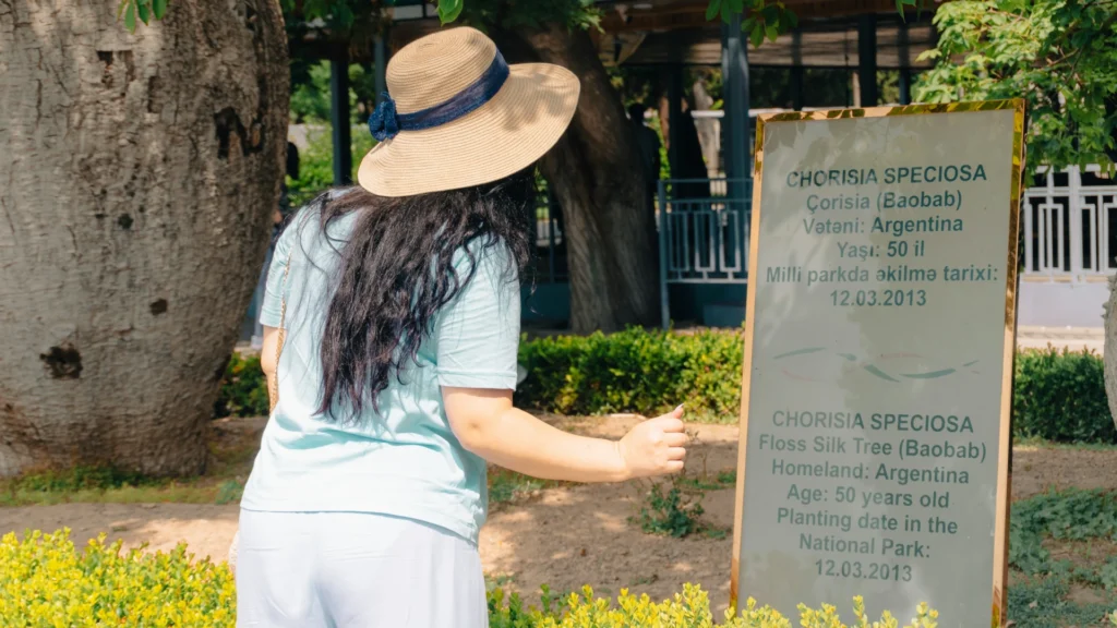 Eine Frau mit langen braunen Haaren steht mit dem Rücken zur Kamera vor einer Infotafel in einem botanischen Garten. Sie trägt einen Strohhut und hellblaue Kleidung.