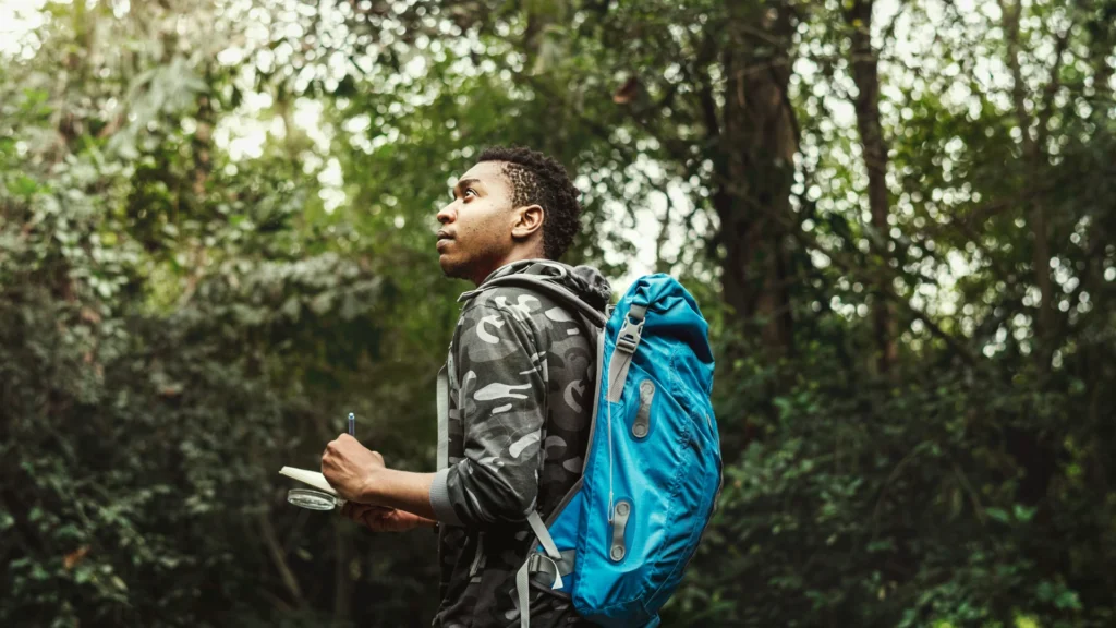 Ein Mann mit kurzen dunklen Haaren, grauer Flecktarnjacke und hellblauem Ruchsack steht in einem Wald. Er schaut aufmerksam und macht sich Notizen.